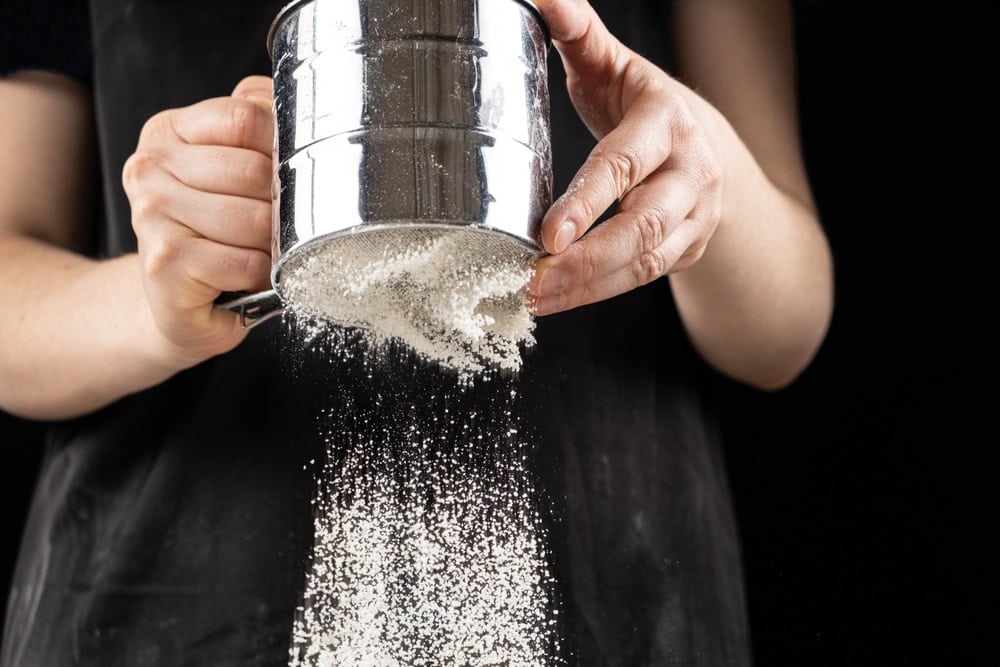 A sifter with flour being sifted into a bowl to remove lumps.